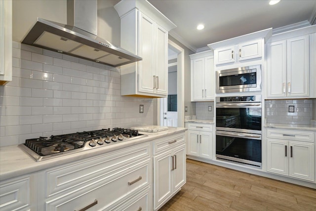 kitchen with light hardwood / wood-style flooring, ornamental molding, white cabinets, appliances with stainless steel finishes, and wall chimney range hood