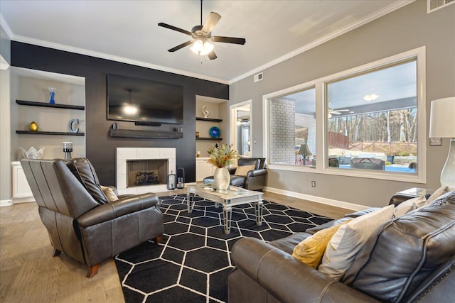 living room with light wood-type flooring, a tiled fireplace, built in shelves, ornamental molding, and ceiling fan