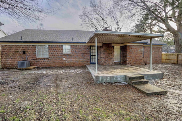 back of property at dusk featuring a deck, cooling unit, brick siding, fence, and roof with shingles