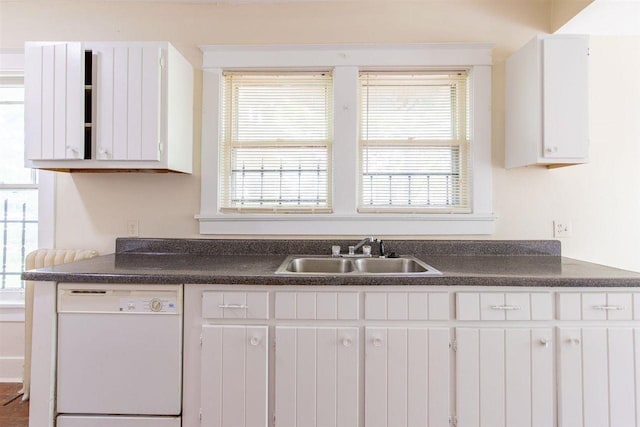 kitchen featuring white dishwasher, a sink, white cabinetry, a healthy amount of sunlight, and dark countertops