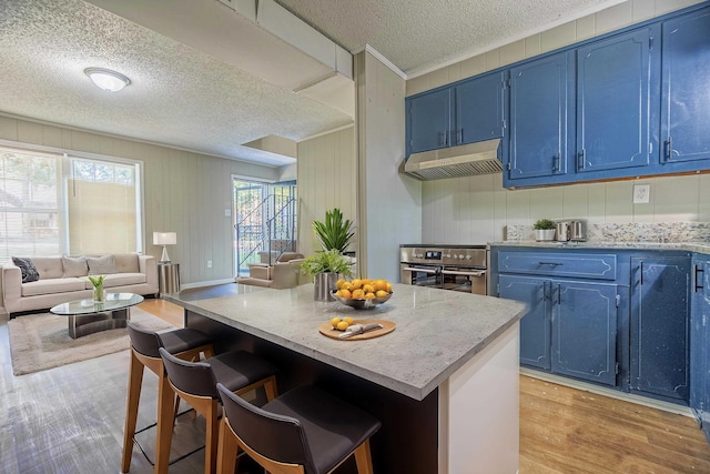 kitchen with blue cabinetry, open floor plan, stainless steel oven, under cabinet range hood, and a kitchen bar