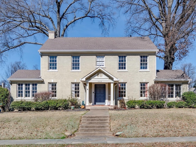 view of front facade with brick siding, a chimney, a front yard, and a shingled roof