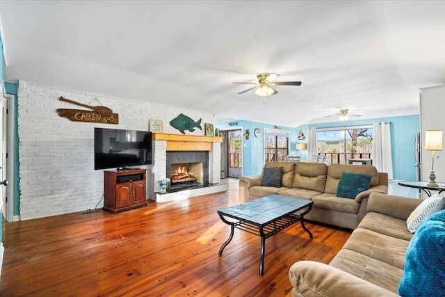 living area with a ceiling fan, a warm lit fireplace, brick wall, and wood finished floors