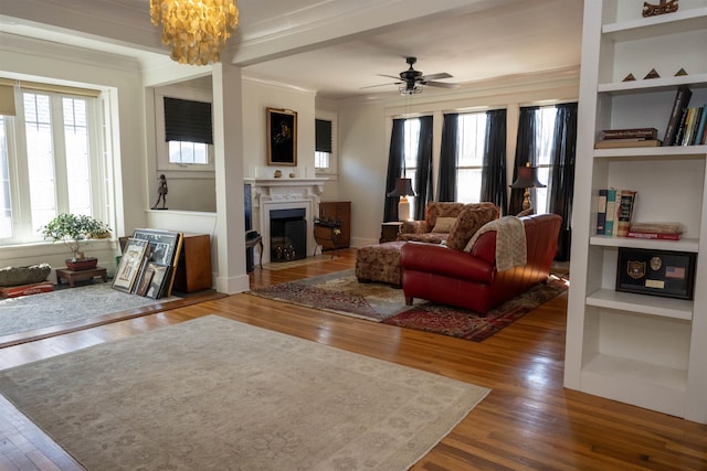 living room with built in features, ornamental molding, a fireplace with flush hearth, wood finished floors, and ceiling fan with notable chandelier