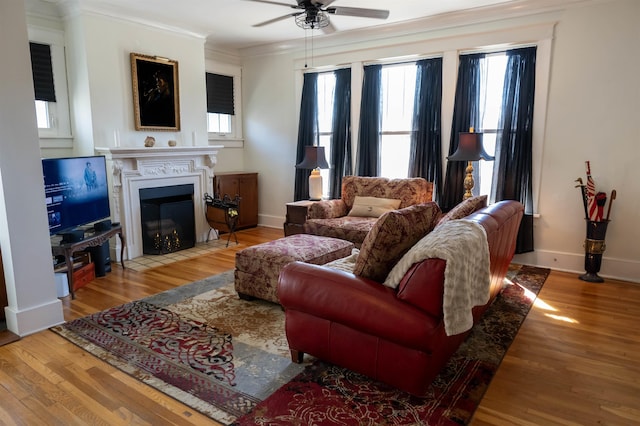 living area featuring baseboards, a fireplace with flush hearth, ceiling fan, wood finished floors, and crown molding