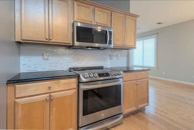 kitchen with backsplash, appliances with stainless steel finishes, dark stone countertops, light wood-type flooring, and baseboards