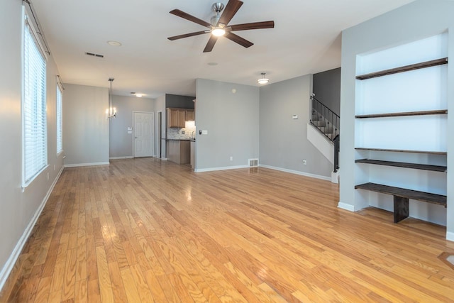 unfurnished living room with light wood-style flooring, visible vents, stairway, and ceiling fan with notable chandelier