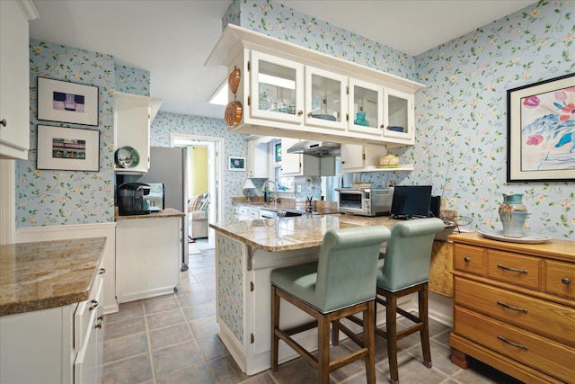 kitchen featuring light stone counters, under cabinet range hood, white cabinetry, glass insert cabinets, and wallpapered walls