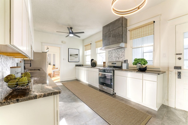 kitchen featuring stainless steel gas range oven, a ceiling fan, white cabinetry, decorative backsplash, and custom range hood