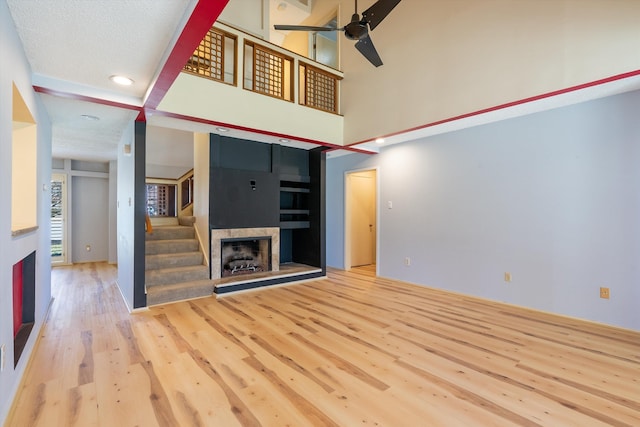 unfurnished living room featuring a fireplace with raised hearth, ceiling fan, a high ceiling, wood finished floors, and stairs