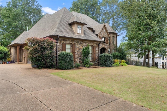 french country inspired facade featuring a shingled roof, concrete driveway, a front yard, a garage, and stone siding