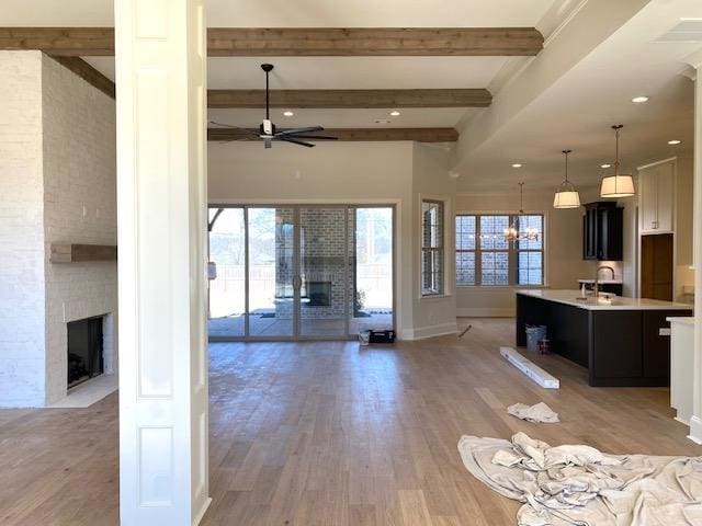 unfurnished living room with a large fireplace, light wood-style flooring, ceiling fan with notable chandelier, and beam ceiling