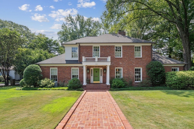 georgian-style home with a chimney, brick siding, a balcony, and a front lawn