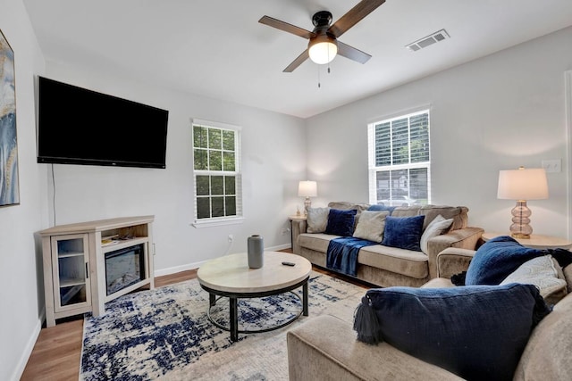 living room with ceiling fan, wood finished floors, visible vents, baseboards, and a glass covered fireplace