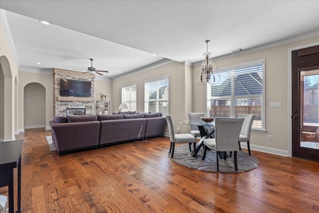 dining room featuring ceiling fan with notable chandelier, arched walkways, ornamental molding, a stone fireplace, and hardwood / wood-style floors