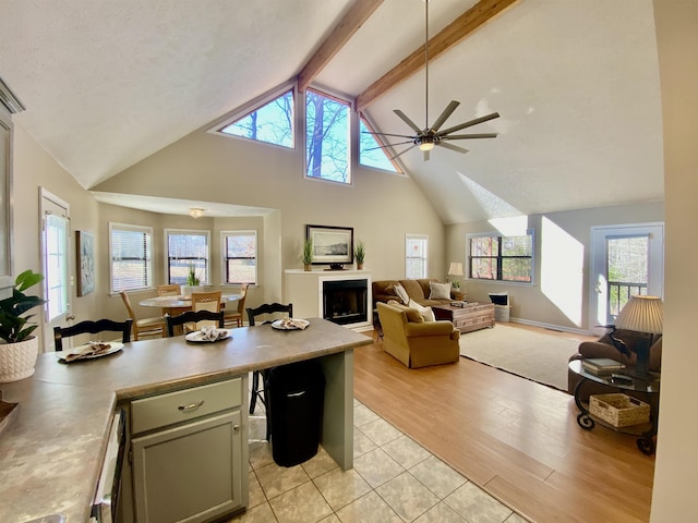 living area with high vaulted ceiling, light wood-style flooring, a fireplace, a ceiling fan, and beam ceiling