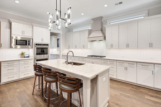 kitchen featuring a kitchen island with sink, premium range hood, a sink, appliances with stainless steel finishes, and light wood-type flooring