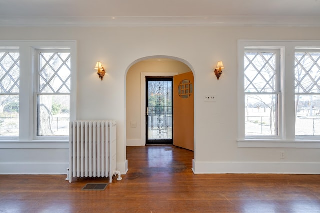 foyer entrance featuring arched walkways, wood finished floors, ornamental molding, a wealth of natural light, and radiator heating unit