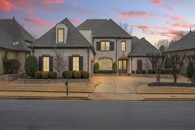 french country home with brick siding, driveway, and roof with shingles