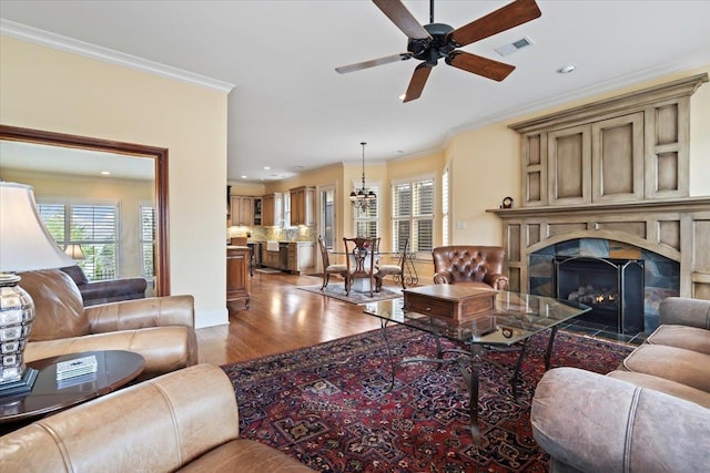 living room featuring a warm lit fireplace, visible vents, ceiling fan, ornamental molding, and wood finished floors