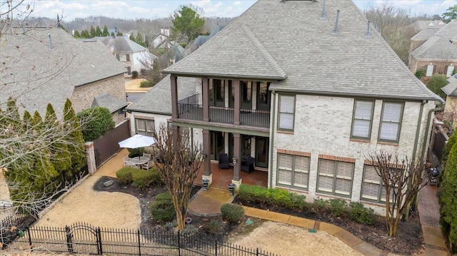 rear view of property with a fenced front yard, roof with shingles, a patio, brick siding, and a balcony