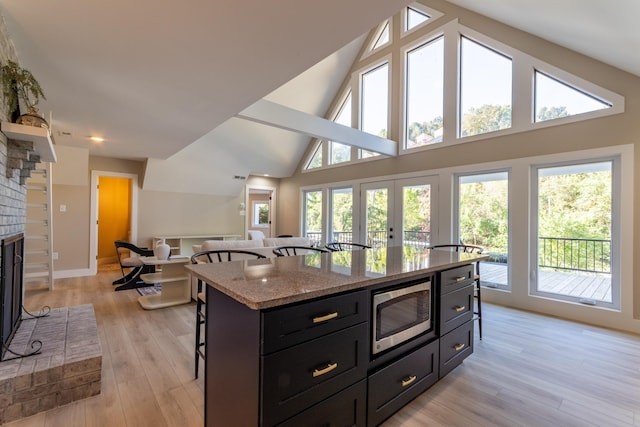 kitchen with light wood-style floors, stainless steel microwave, dark cabinetry, a brick fireplace, and a kitchen bar