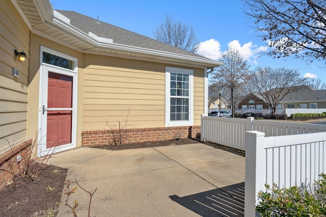 view of exterior entry featuring a patio, a shingled roof, fence, and brick siding