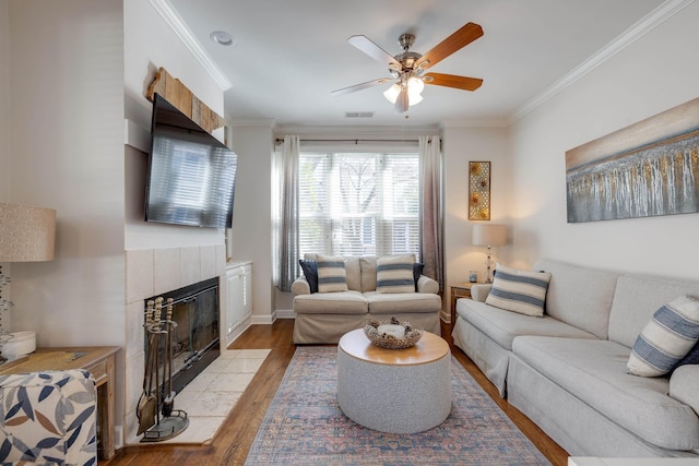 living area with visible vents, a ceiling fan, a tiled fireplace, light wood-style flooring, and crown molding