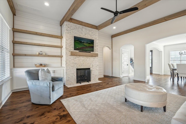 living area featuring arched walkways, ceiling fan, a stone fireplace, dark wood-type flooring, and beamed ceiling