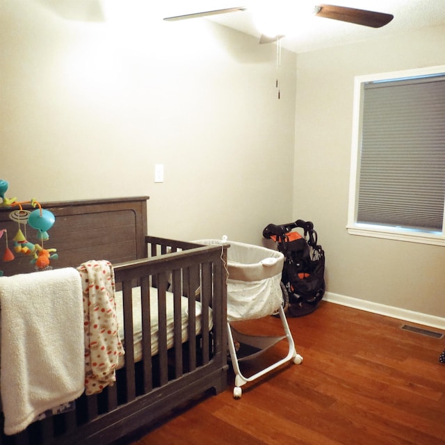 bedroom featuring a crib, baseboards, visible vents, ceiling fan, and wood finished floors