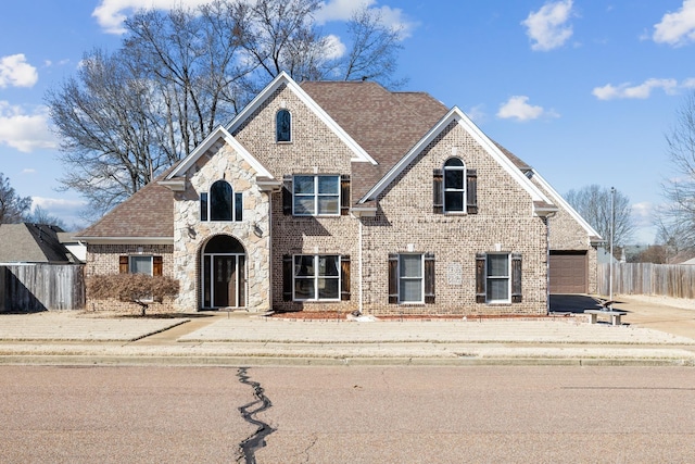 traditional home featuring stone siding, brick siding, fence, and roof with shingles