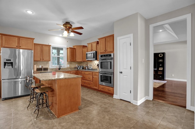 kitchen featuring a center island, stainless steel appliances, a sink, ceiling fan, and light stone countertops