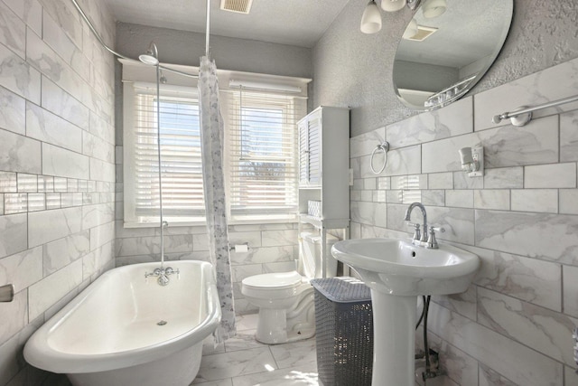 full bath featuring marble finish floor, tile walls, a textured wall, toilet, and a freestanding tub