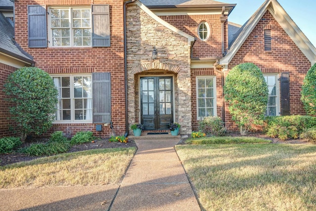 view of exterior entry featuring french doors, brick siding, a yard, a shingled roof, and stone siding