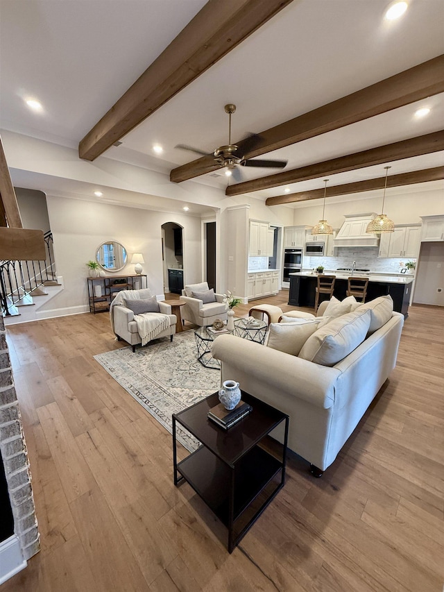 living room with beam ceiling, light wood-style floors, a ceiling fan, baseboards, and stairs