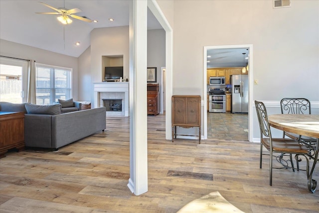 living area featuring high vaulted ceiling, a fireplace, visible vents, a ceiling fan, and light wood finished floors