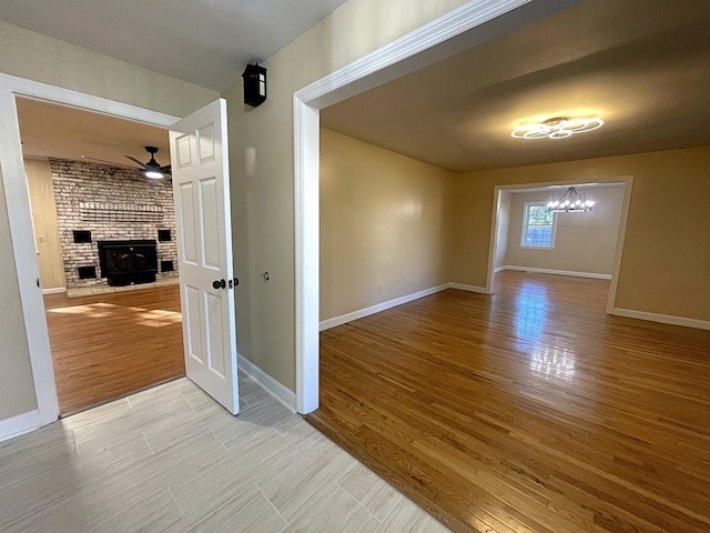 spare room with ceiling fan with notable chandelier, a brick fireplace, wood finished floors, and baseboards