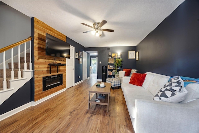 living room with stairway, a glass covered fireplace, ceiling fan, wood finished floors, and baseboards