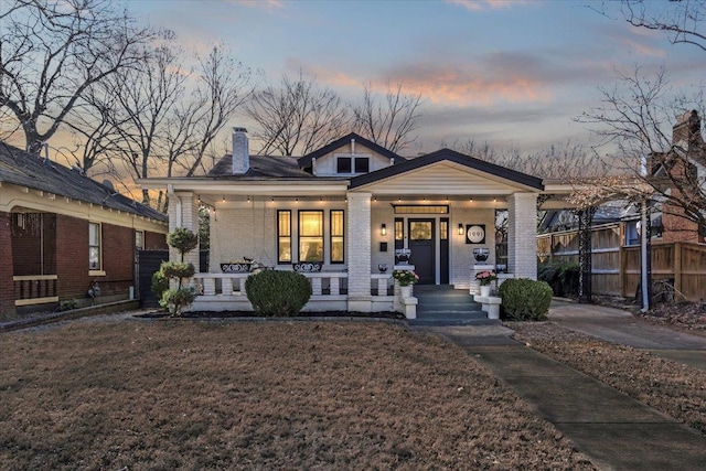 view of front of house with brick siding, a chimney, covered porch, fence, and a front lawn