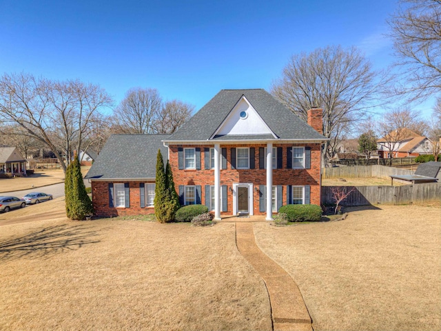 greek revival house with brick siding, a shingled roof, a chimney, fence, and a front yard