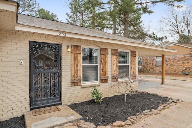 view of exterior entry with driveway, a carport, roof with shingles, and brick siding