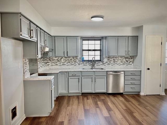 kitchen featuring under cabinet range hood, gray cabinetry, dark wood-type flooring, a sink, and stainless steel dishwasher