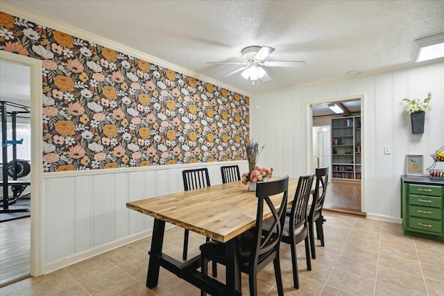 dining space featuring light tile patterned floors, a wainscoted wall, ceiling fan, ornamental molding, and a textured ceiling