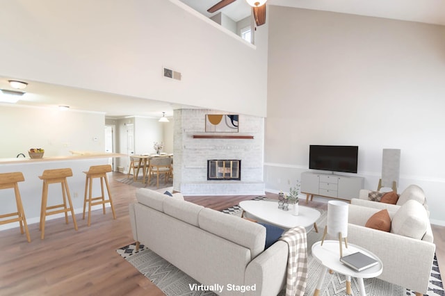 living area with visible vents, a towering ceiling, a ceiling fan, a brick fireplace, and wood finished floors