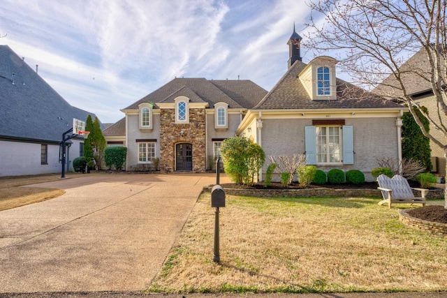 french provincial home featuring brick siding, a shingled roof, concrete driveway, a chimney, and a front yard