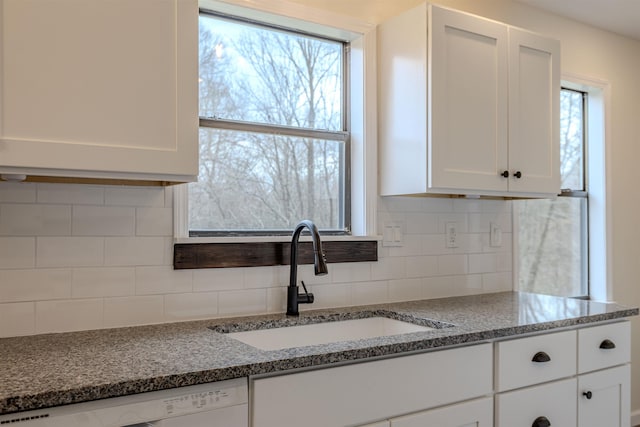 kitchen featuring decorative backsplash, dark stone countertops, white dishwasher, white cabinetry, and a sink