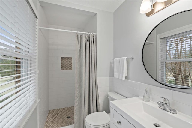 full bathroom with toilet, a wainscoted wall, a tile shower, vanity, and tile walls