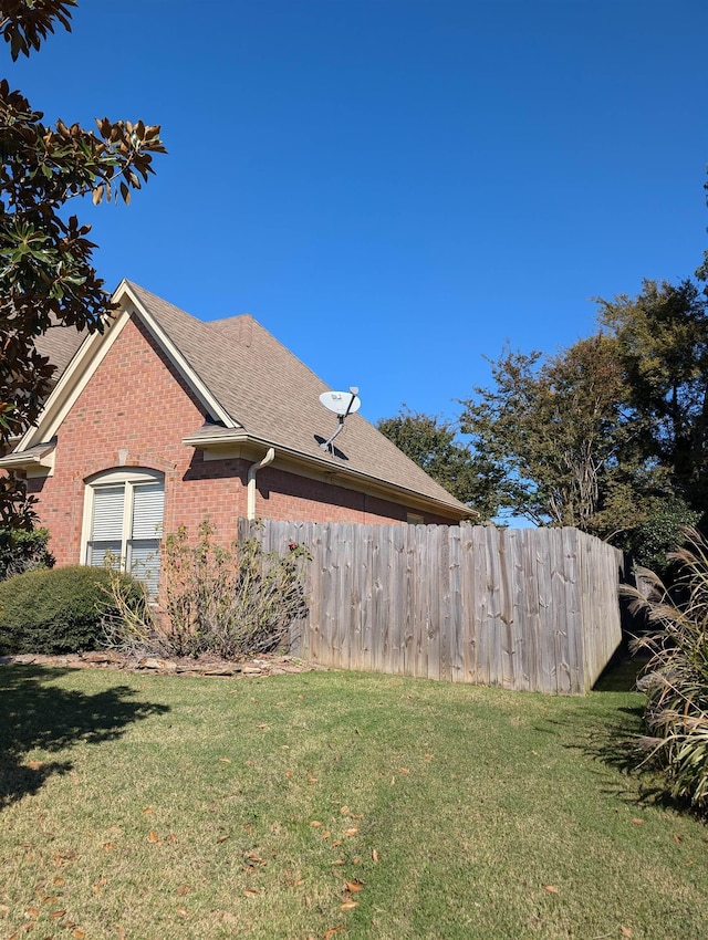 view of side of home with roof with shingles, fence, a lawn, and brick siding