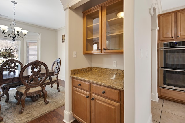 kitchen featuring double oven, glass insert cabinets, ornamental molding, light stone countertops, and a chandelier