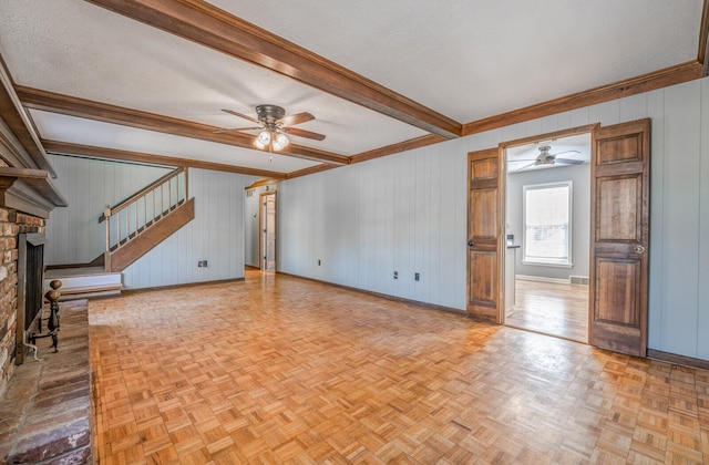 unfurnished living room with a brick fireplace, ceiling fan, beamed ceiling, baseboards, and stairs